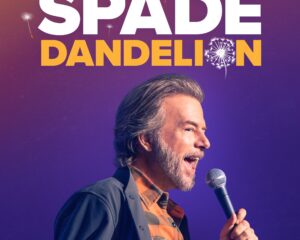 Comedian David Spade performing on stage with a microphone, against a purple-orange gradient poster that reads 'DAVID SPADE DANDELION'.