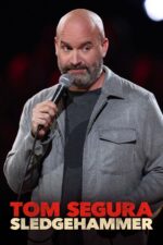 Comedian on stage holding a microphone, wearing a gray zip‑front jacket over a dark shirt; bold red and white text at the bottom reads 'TOM SEGURA SLEDGEHAMMER'