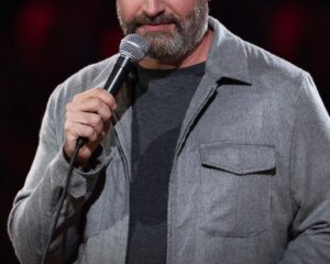 Comedian on stage holding a microphone, wearing a gray zip‑front jacket over a dark shirt; bold red and white text at the bottom reads 'TOM SEGURA SLEDGEHAMMER'