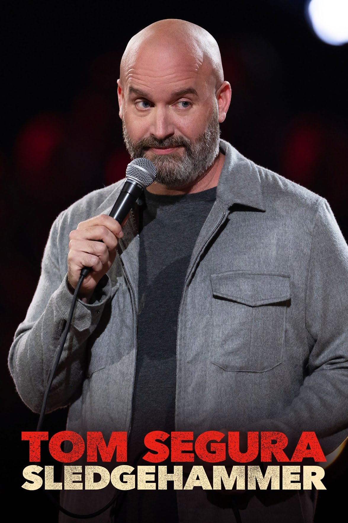 Comedian on stage holding a microphone, wearing a gray zip‑front jacket over a dark shirt; bold red and white text at the bottom reads 'TOM SEGURA SLEDGEHAMMER'