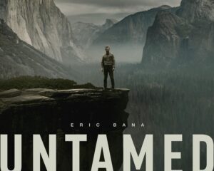 Man stands on a cliff edge overlooking a misty canyon with towering rock faces in a dramatic mountain landscape, movie poster for Untamed featuring Eric Bana.