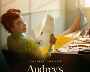 Poster for Audrey’s Children featuring a retro-looking woman reading a document at a cluttered desk bathed in warm sunlight; title and credits at bottom.
