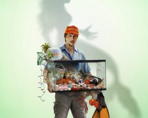 Man in cap carries a glass display case brimming with toys and souvenirs on a sandy beach, with a sandcastle nearby and a bright green backdrop.